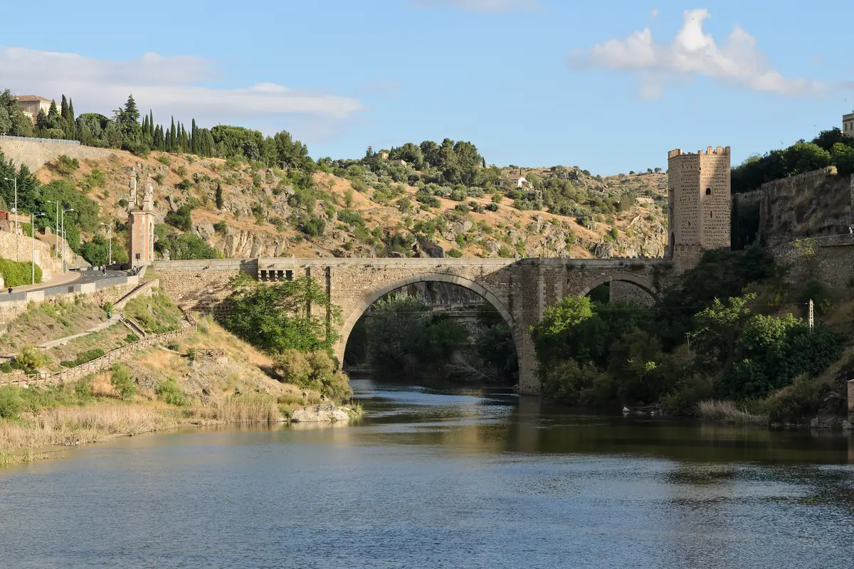 The stone Alcántara Bridge over the Tagus, with a gate tower at its right end and dry, wooded slopes rising behind the river