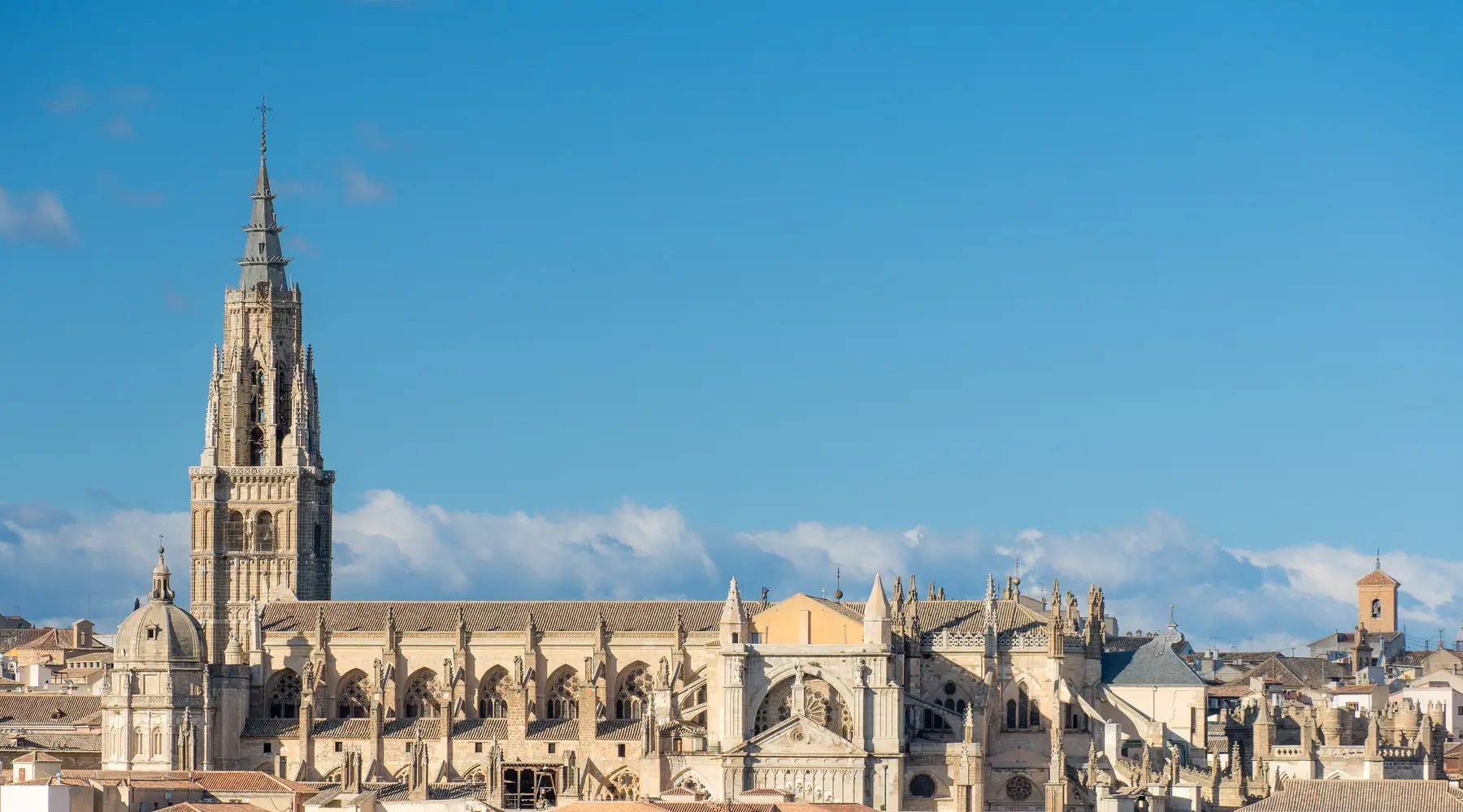 Toledo Cathedral's tall Gothic spire and long stone roofline above a sea of terracotta rooftops under a blue sky